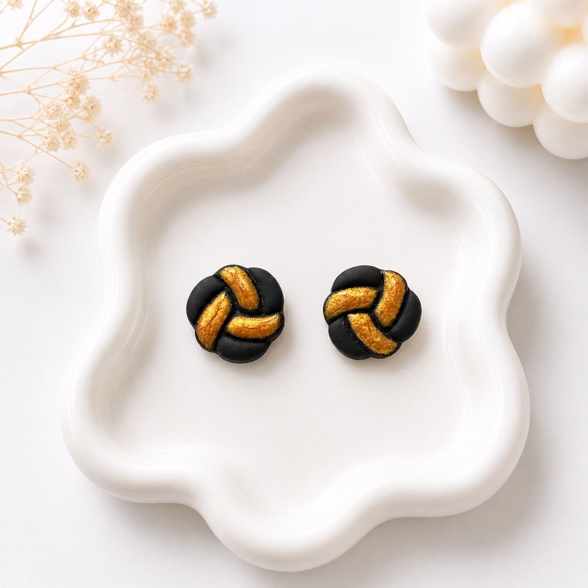A pair of round black clay stud earrings with a braided knot pattern and gold-painted grooves, displayed on a white ceramic tray.