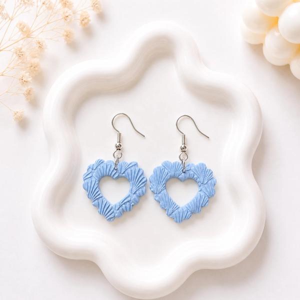 A pair of periwinkle blue clay dangle earrings in an open heart shape covered with seashell relief texture, displayed on a white wavy tray.