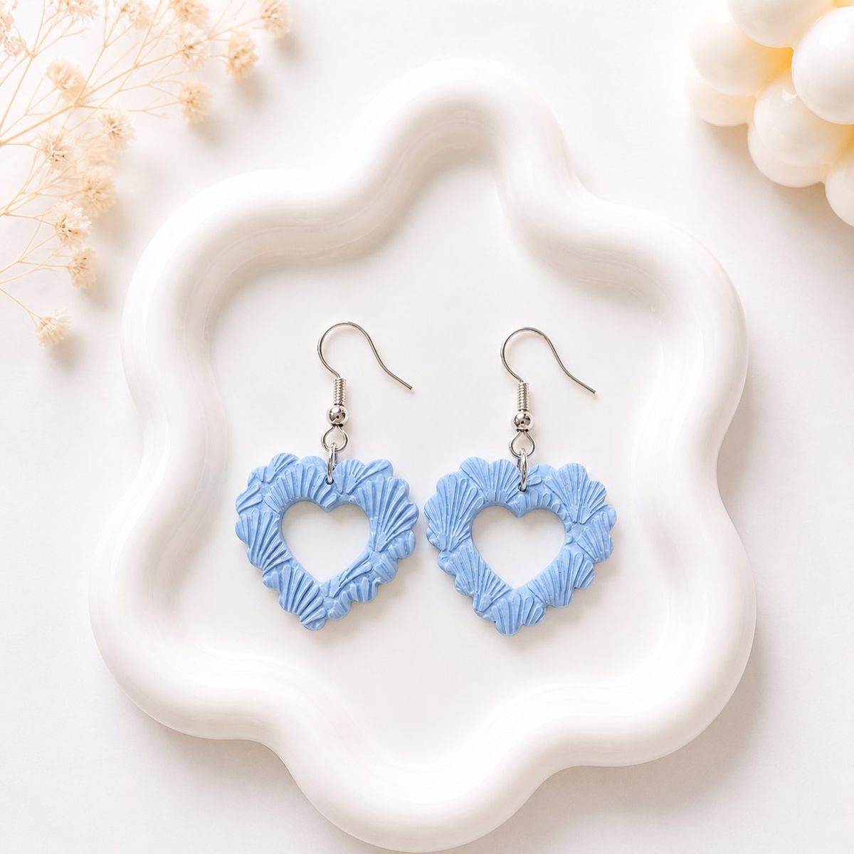A pair of periwinkle blue clay dangle earrings in an open heart shape covered with seashell relief texture, displayed on a white wavy tray.