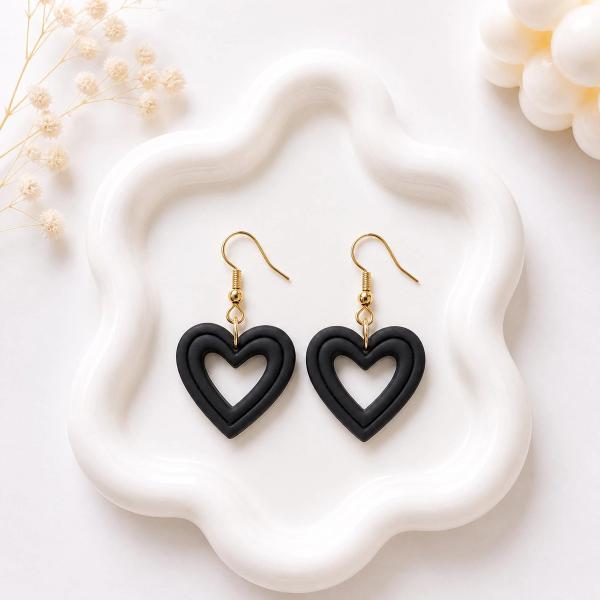 A pair of matte black clay open-heart dangle earrings with gold-tone fish hook findings displayed on a white ceramic tray.