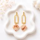 A pair of dangle earrings with gold arch connectors and ball studs, hanging square clay pendants in a beige and brown marble pattern, displayed on a white ceramic tray.
