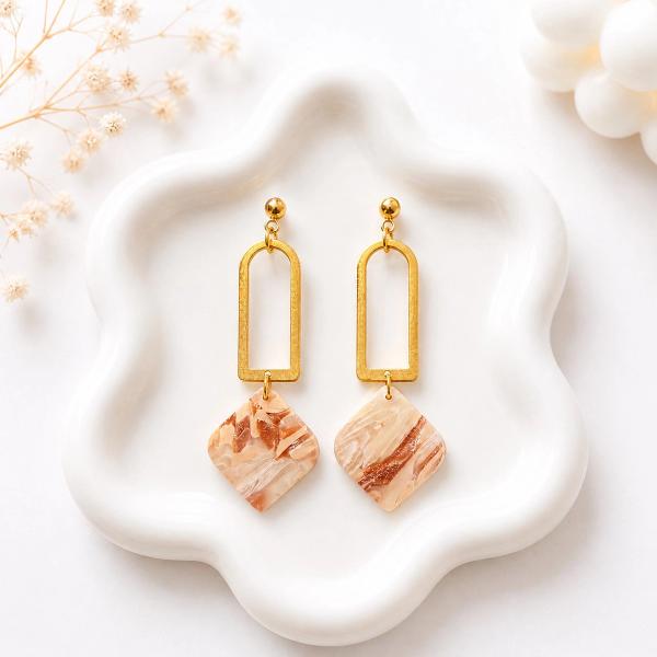 A pair of dangle earrings with gold arch connectors and ball studs, hanging square clay pendants in a beige and brown marble pattern, displayed on a white ceramic tray.