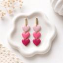 A pair of dangle earrings with three stacked clay hearts in graduating shades of pink attached to gold-tone bar studs, displayed on a white ceramic dish.