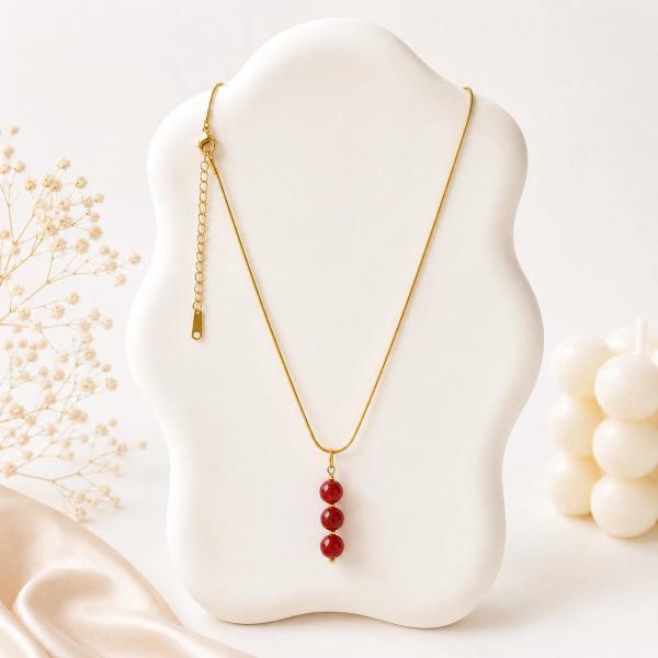 A gold snake chain necklace with three graduating deep red round bead pendants displayed on a white jewelry bust.