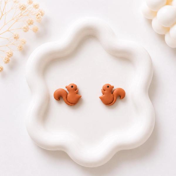 A pair of small terracotta brown clay squirrel stud earrings displayed on a white ceramic tray.