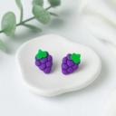 A pair of purple polymer clay grape cluster stud earrings with green leaf tops resting on a white ceramic dish.