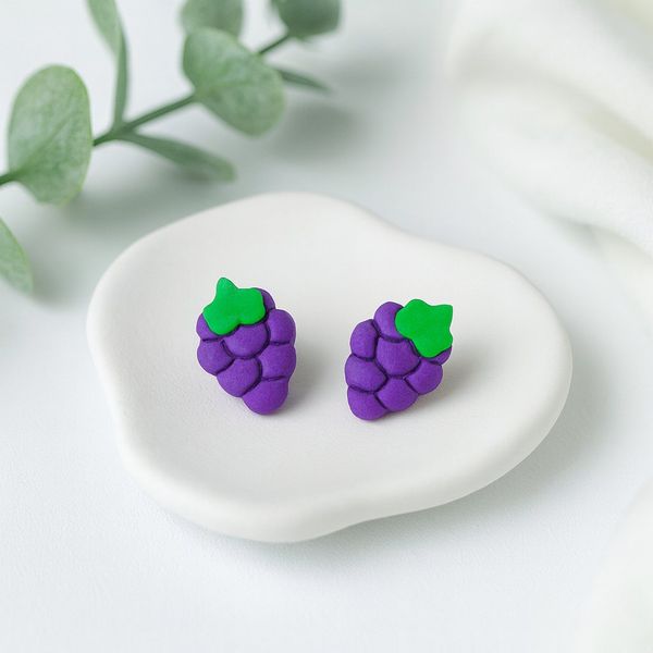 A pair of purple polymer clay grape cluster stud earrings with green leaf tops resting on a white ceramic dish.