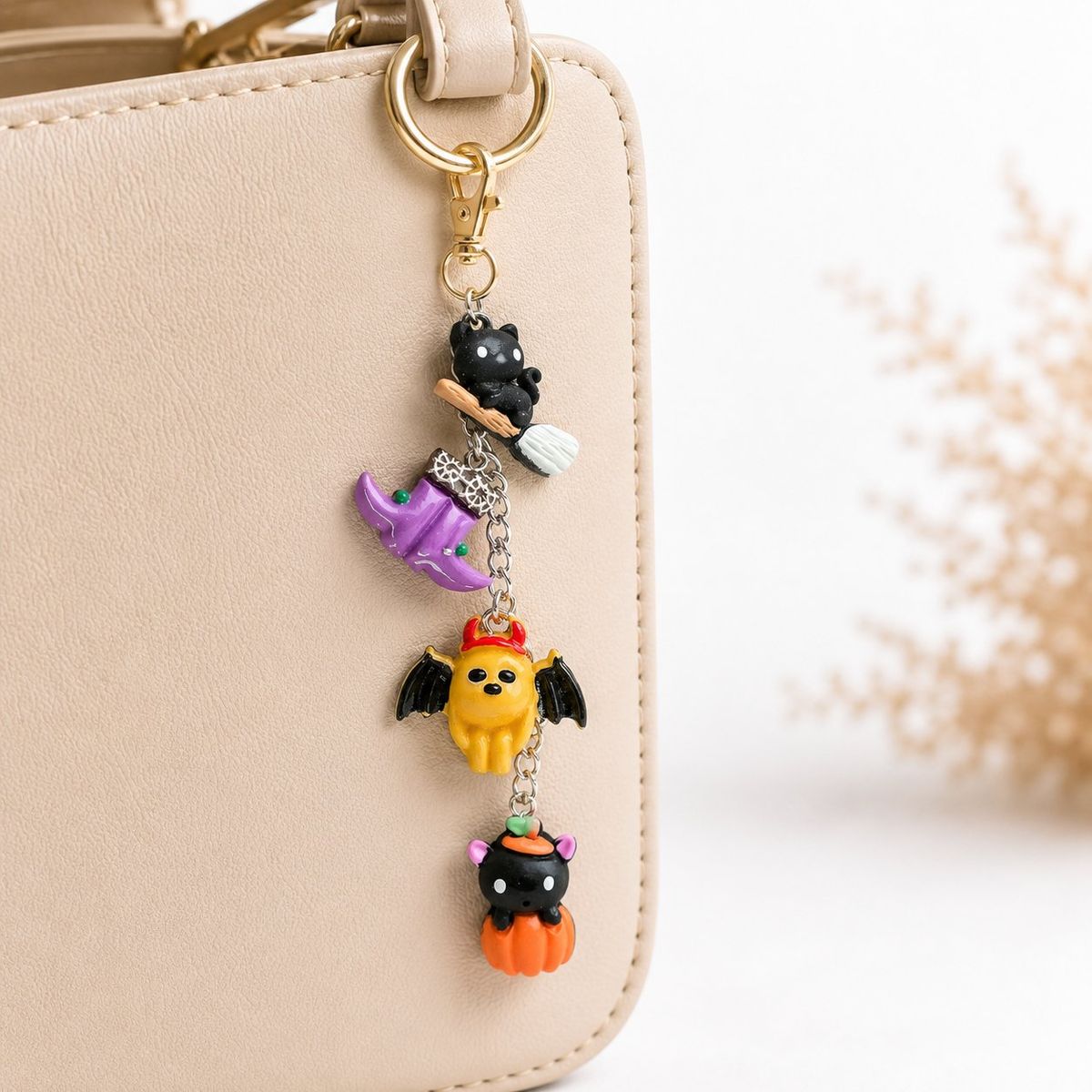 A Halloween-themed keychain charm with multiple polymer clay figures — a black cat on a broomstick, a purple witch boot, a yellow devil-bat creature, and a black cauldron on an orange pumpkin — connected by silver chain links with a gold lobster clasp, clipped to a beige leather bag.