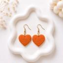 A pair of orange clay heart-shaped dangle earrings with an embossed fan texture and gold-toned hook findings, displayed on a white wavy tray.