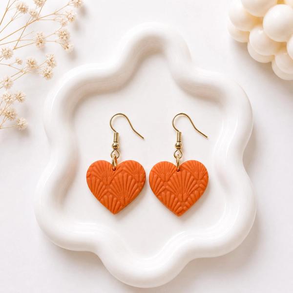 A pair of orange clay heart-shaped dangle earrings with an embossed fan texture and gold-toned hook findings, displayed on a white wavy tray.
