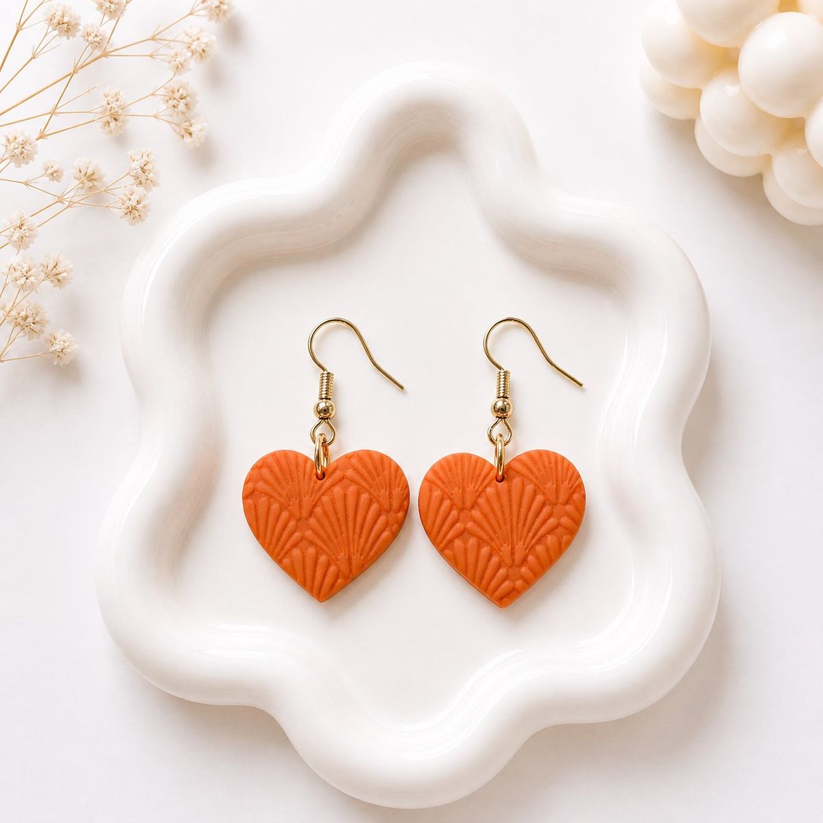 A pair of orange clay heart-shaped dangle earrings with an embossed fan texture and gold-toned hook findings, displayed on a white wavy tray.