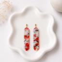 A pair of elongated bar dangle earrings with a red, white, and black marble swirl clay pattern, hanging from small gold ball studs, displayed on a white ceramic tray.