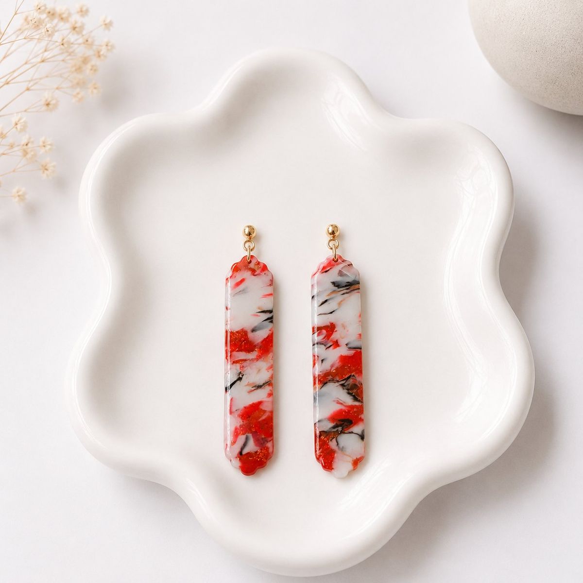 A pair of elongated bar dangle earrings with a red, white, and black marble swirl clay pattern, hanging from small gold ball studs, displayed on a white ceramic tray.