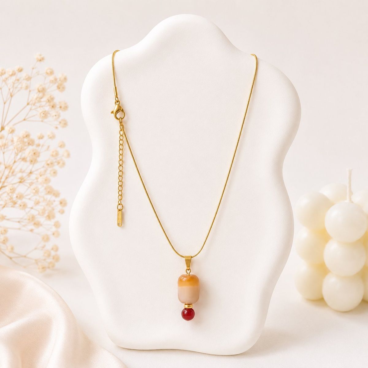 A gold snake chain necklace displayed on a white bust, featuring an amber cylindrical bead pendant with a small red bead accent below it.