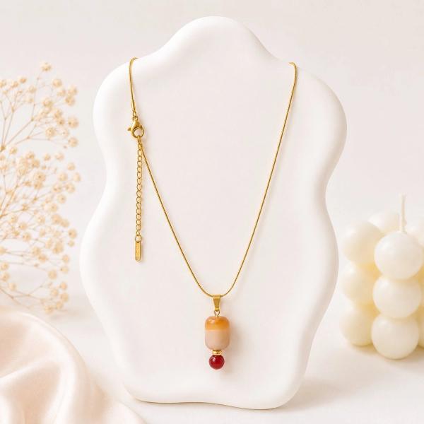 A gold snake chain necklace displayed on a white bust, featuring an amber cylindrical bead pendant with a small red bead accent below it.