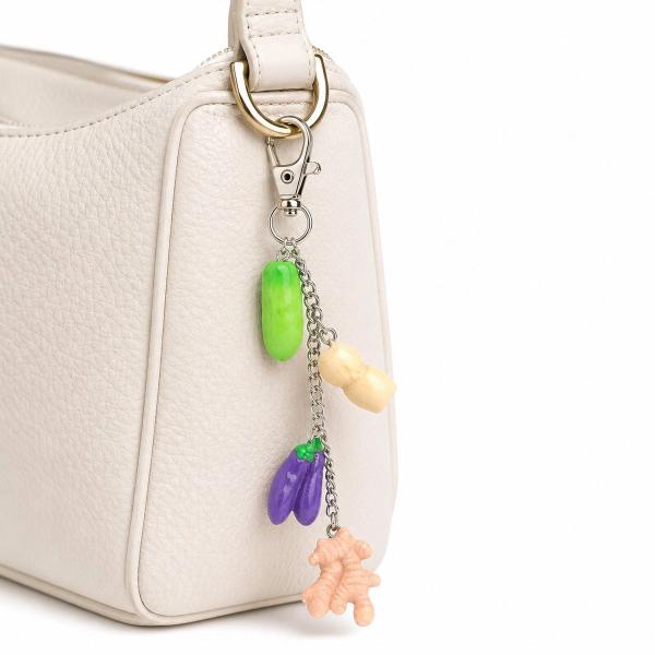 A key charm with four polymer clay vegetable charms — green cucumber, cream garlic, purple eggplant, and peach coral root — hanging from a silver chain clipped onto a cream leather handbag.