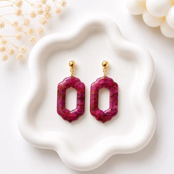 A pair of deep burgundy marbled clay dangle earrings with gold glitter accents and an open hexagonal frame, hanging from gold ball studs on a white ceramic dish.