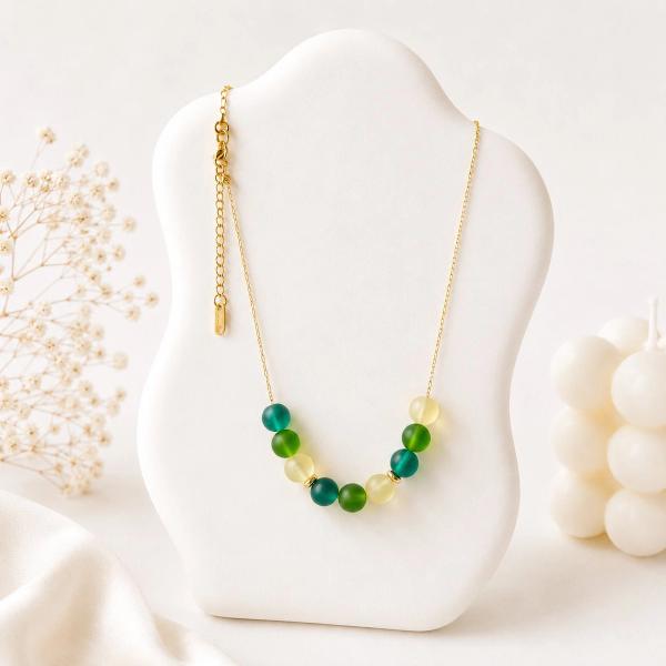 A gold chain necklace with a curved row of round green and yellow-green beads in frosted and glossy finishes displayed on a white jewelry bust.