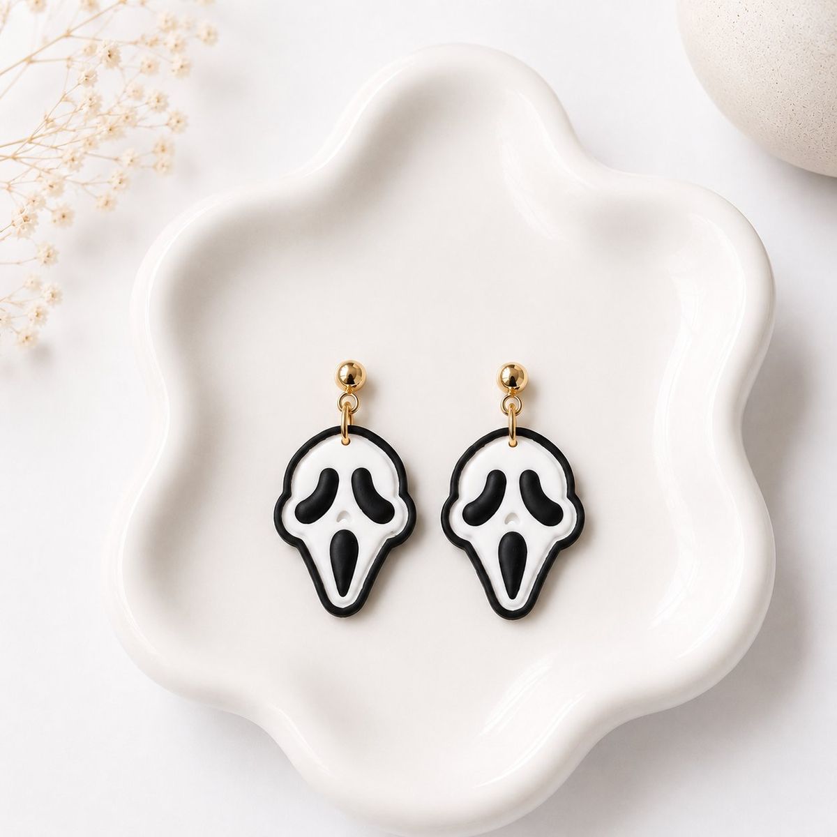 A pair of black and white clay ghost face mask dangle earrings with gold ball stud tops, displayed on a white ceramic dish.