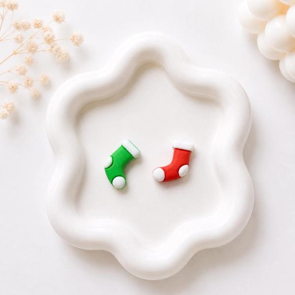 Two small polymer clay stud earrings shaped like Christmas stockings, one green and one red, each with white trim and dot details, displayed on a white wavy tray.