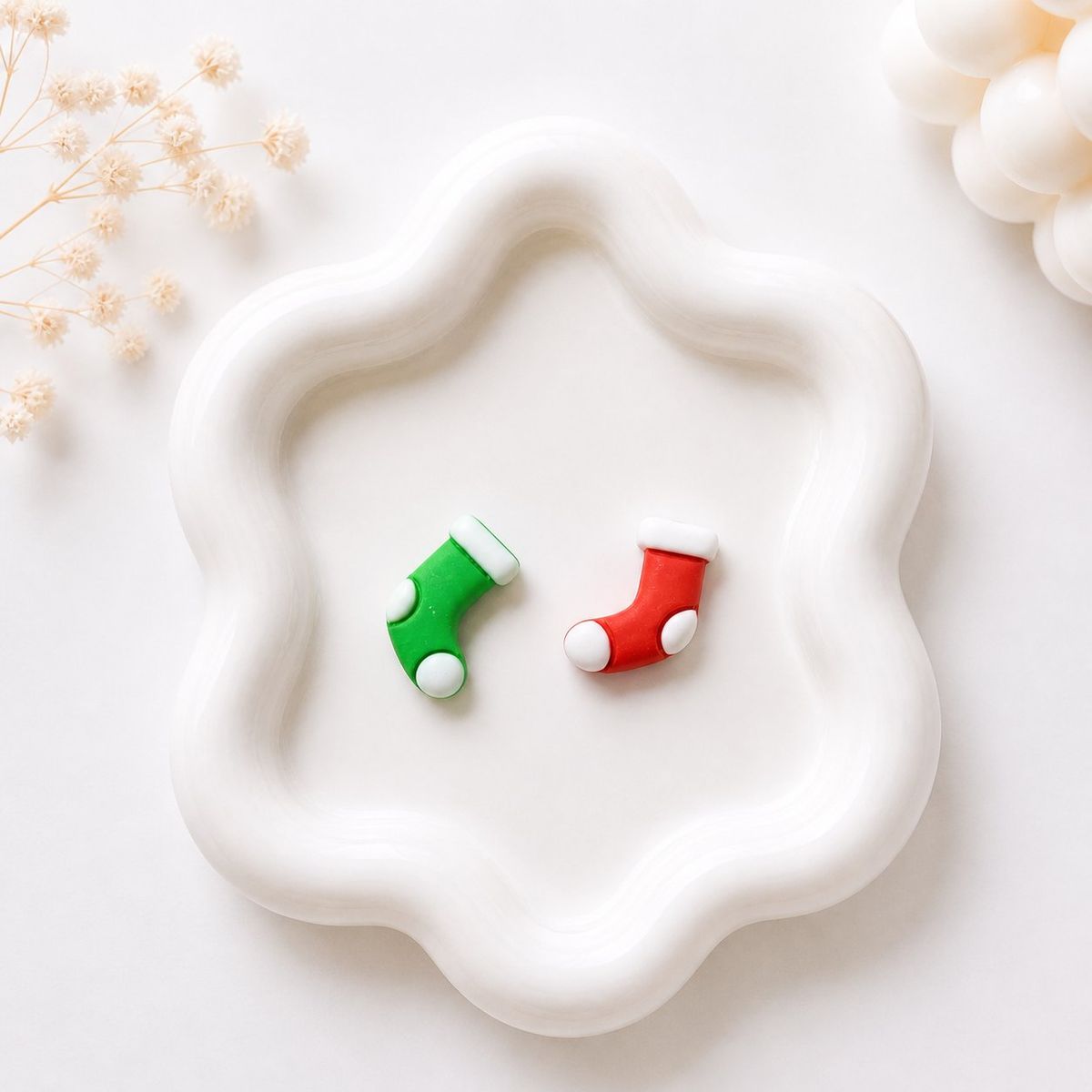 Two small polymer clay stud earrings shaped like Christmas stockings, one green and one red, each with white trim and dot details, displayed on a white wavy tray.