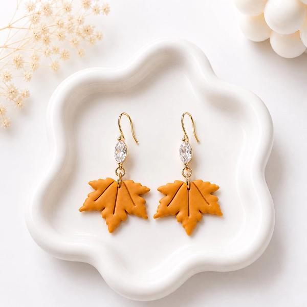 A pair of orange polymer clay maple leaf dangle earrings with clear marquise crystal connectors and gold-toned hooks, displayed on a white ceramic tray.