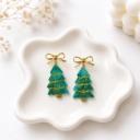 A pair of green resin Christmas tree dangle earrings with gold glitter detailing, hanging from gold metal bow studs, displayed on a white ceramic dish.