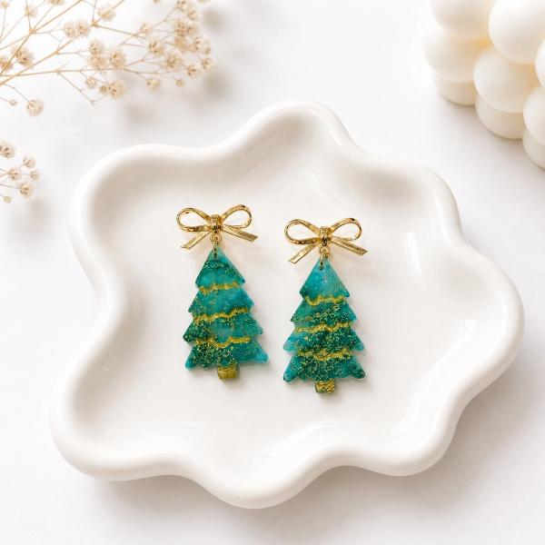 A pair of green resin Christmas tree dangle earrings with gold glitter detailing, hanging from gold metal bow studs, displayed on a white ceramic dish.