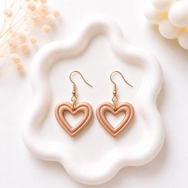 A pair of open heart-shaped rose gold shimmer clay dangle earrings with gold fish hook wires, displayed on a white ceramic tray.