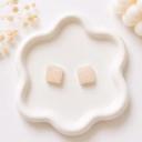 A pair of small square cream-colored stud earrings with iridescent glitter detail, displayed on a white wavy ceramic tray.
