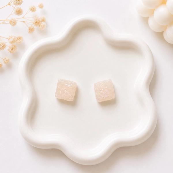 A pair of small square cream-colored stud earrings with iridescent glitter detail, displayed on a white wavy ceramic tray.