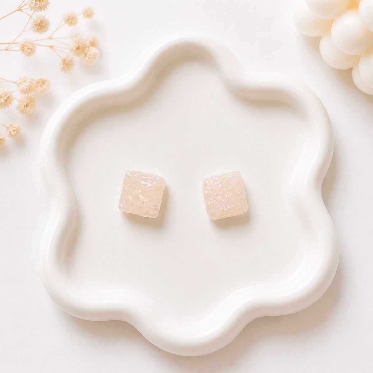 A pair of small square cream-colored stud earrings with iridescent glitter detail, displayed on a white wavy ceramic tray.