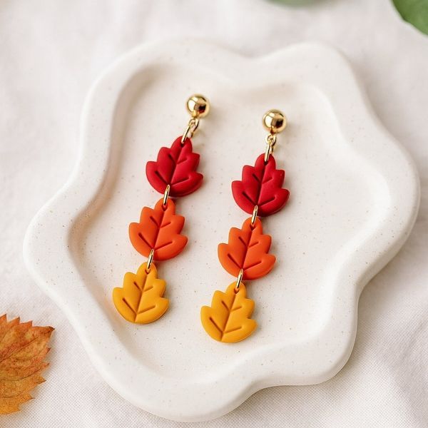 Three-tiered clay leaf dangle earrings in red, orange, and yellow with gold ball studs displayed on a white ceramic dish.