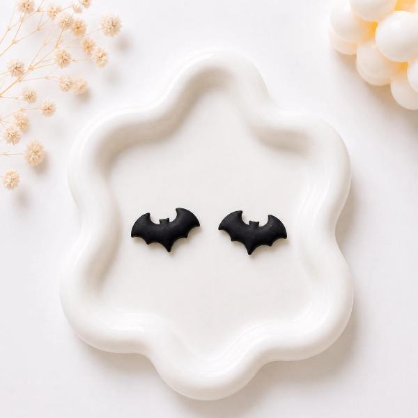 A pair of small matte black bat-shaped clay stud earrings displayed on a white ceramic tray.