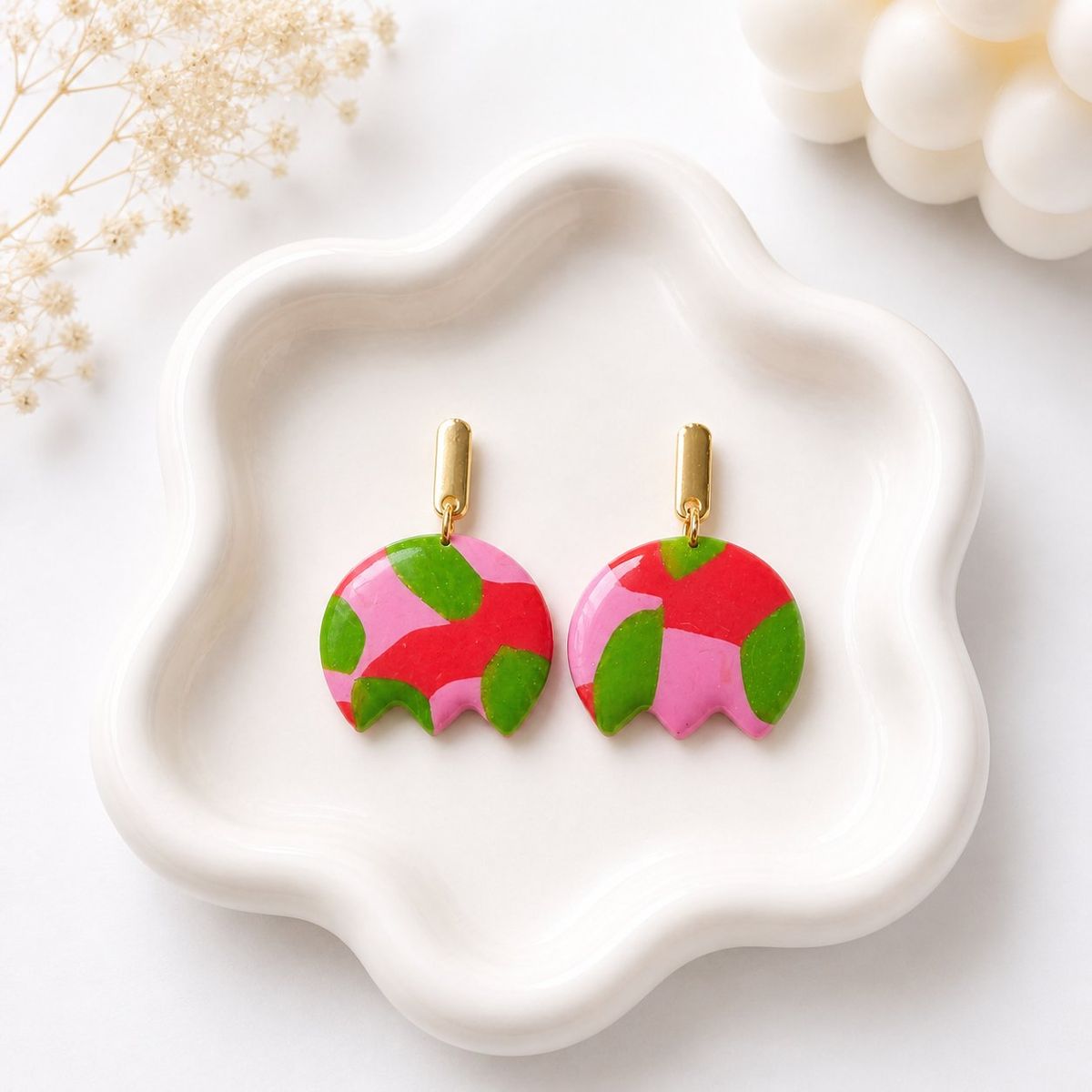 A pair of round clay dangle earrings with a pink, red, and green abstract floral pattern and scalloped bottom edge, hanging from gold bar studs, displayed on a white ceramic tray.