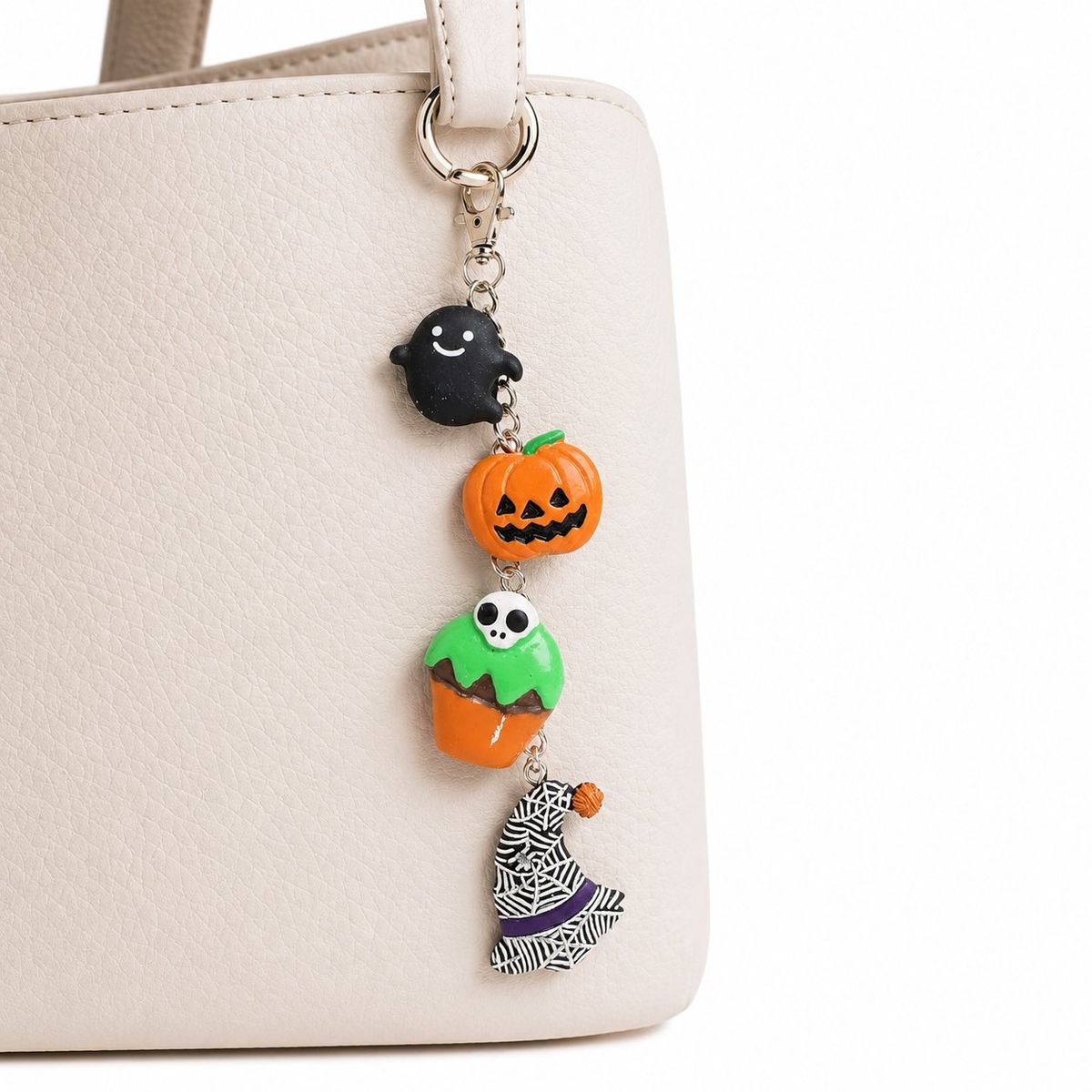 A Halloween-themed bag charm with four charms — a black ghost, orange jack-o'-lantern, green skull cupcake, and a witch figure with spider web print — attached to a cream leather handbag via silver lobster clasp.