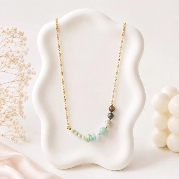 A gold-tone chain necklace displayed on a white bust featuring mint green chip stones, a pale blue round bead, a speckled bead, and two dark grey beads arranged in a curved bar style.