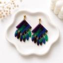 A pair of handmade polymer clay dangle earrings with a marbled blue, green, and purple diamond-shaped top and multiple oval fringe drops attached with gold jump rings, displayed on a white ceramic dish.