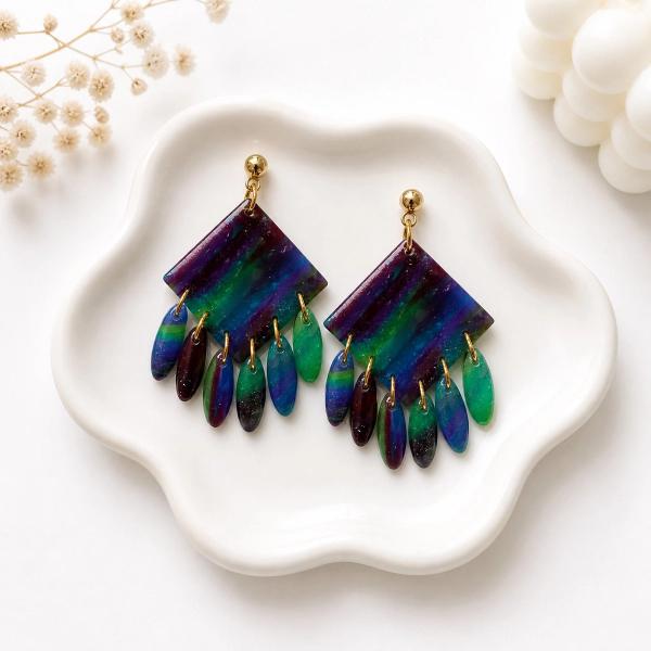 A pair of handmade polymer clay dangle earrings with a marbled blue, green, and purple diamond-shaped top and multiple oval fringe drops attached with gold jump rings, displayed on a white ceramic dish.