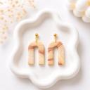 A pair of U-shaped clay dangle earrings with a beige and terracotta marble swirl pattern, attached to gold ball studs, displayed on a white ceramic tray.