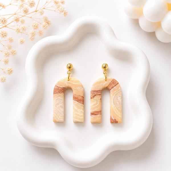 A pair of U-shaped clay dangle earrings with a beige and terracotta marble swirl pattern, attached to gold ball studs, displayed on a white ceramic tray.