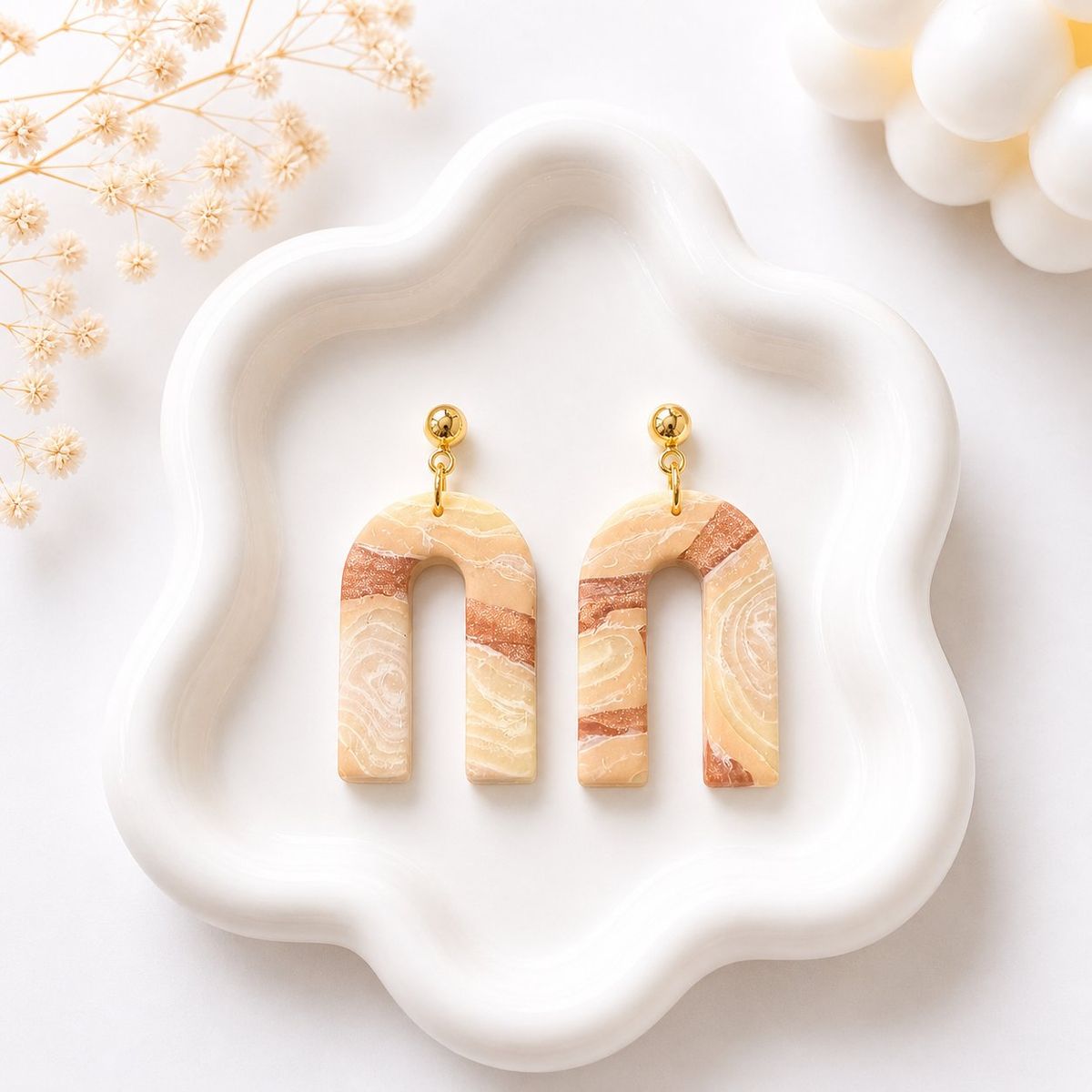 A pair of U-shaped clay dangle earrings with a beige and terracotta marble swirl pattern, attached to gold ball studs, displayed on a white ceramic tray.