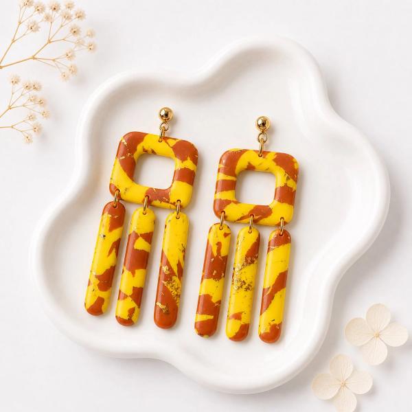 A pair of handmade polymer clay dangle earrings with a yellow and rust brown marbled pattern, featuring an open square frame and three hanging rounded bar charms with gold leaf flecks, displayed on a white ceramic tray.