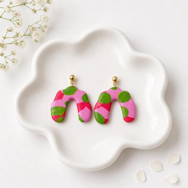 A pair of arch-shaped polymer clay dangle earrings with a pink, red, and green abstract pattern, hung from gold ball studs, displayed on a white ceramic tray.