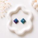 Two small diamond-shaped stud earrings displayed on a white ceramic tray, one with purple and blue marbling and one with teal and green marbling.