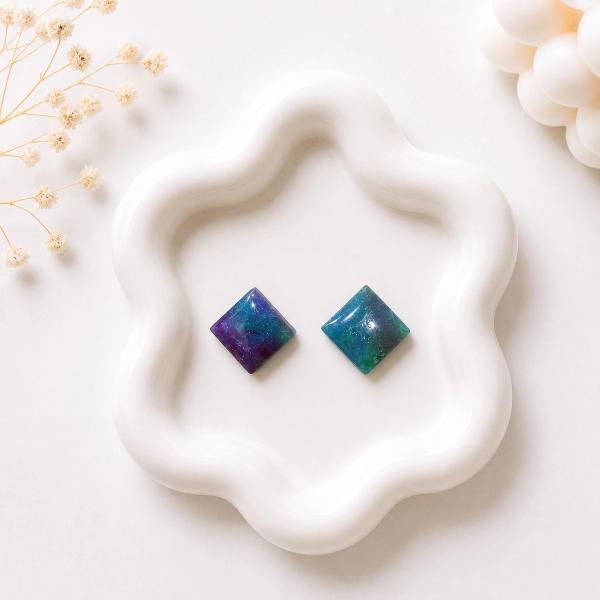 Two small diamond-shaped stud earrings displayed on a white ceramic tray, one with purple and blue marbling and one with teal and green marbling.
