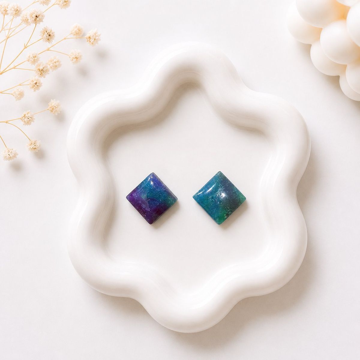 Two small diamond-shaped stud earrings displayed on a white ceramic tray, one with purple and blue marbling and one with teal and green marbling.