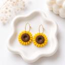 A pair of handmade clay sunflower earrings with yellow petals and brown textured centers hanging from gold hoop wires, displayed on a white ceramic tray.