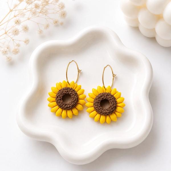 A pair of handmade clay sunflower earrings with yellow petals and brown textured centers hanging from gold hoop wires, displayed on a white ceramic tray.