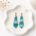 Two elongated pointed clay dangle earrings with teal, mint, and grey swirl patterns hanging from gold ball studs, displayed on a white ceramic dish.